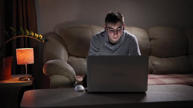 A nerd boy sits on a sofa near window, working on his laptop