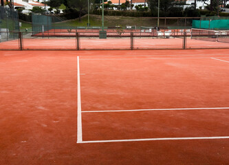 A clay court, marked with bright white lines, awaits the next set. Tennis balls scattered across the surface hint at a recent practice. Greenery surrounds the outdoor space.