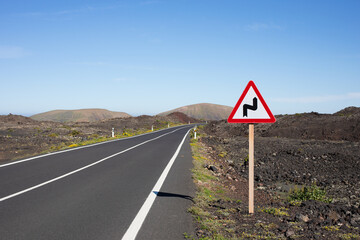 Fototapeta premium A straight, black, endless road with hills and mountains in the background. Spain, Lanzarote. A sign on the side of the road
