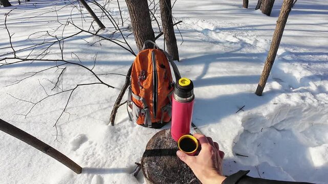 Individual Explorer Displays Cup In Snowy Landscape. Solitary Adventurer Shows Pink Thermos Cup Amidst Winter Woods. Lone Trekker Uncovers Cup From Pink Thermos Beside Orange Bag During Snowy Hike