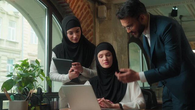 Arabian three coworkers business team in office talking looking laptop teamwork women man Muslim intern in hijab with computer pc job Arabian businessman female secretary coming helping mentor teach