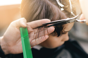 Young caucasian child receiving haircut with scissors and comb in hairstyling salon barbershop, sunlight