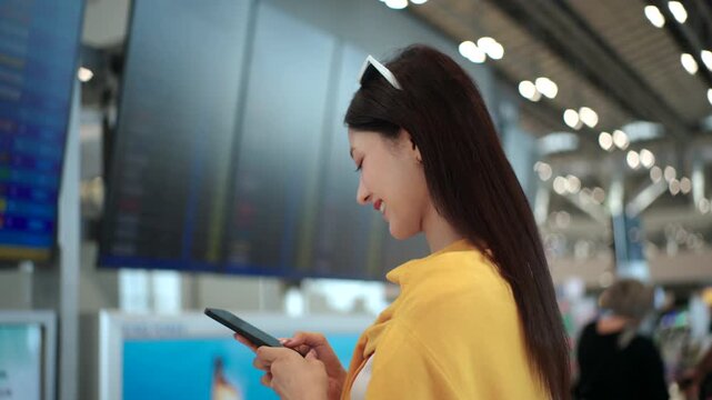 Happy young woman solo traveler using smartphone and holding boarding pass at airport terminal, ready for vacation. Modern travel lifestyle, digital travel planning and travel insurance concept.
