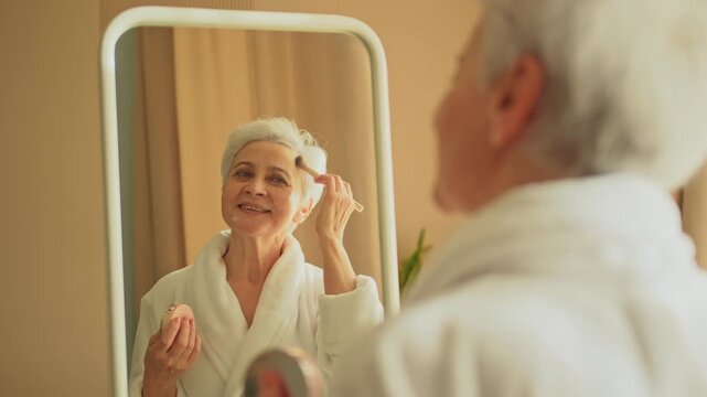 Mature adult old woman with smooth soft perfect skin applying rouge with brush on cheek looking at mirror doing daily beauty make up routine. Senior woman putting blush on her face. Anti age makeup