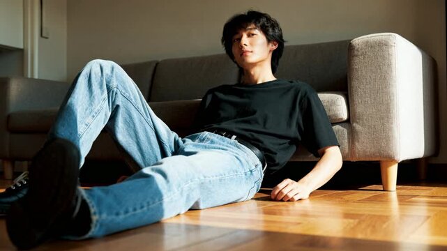 Adult Asian man reclining on parquet by sofa reacting to warming light and shadow, adjusting gaze