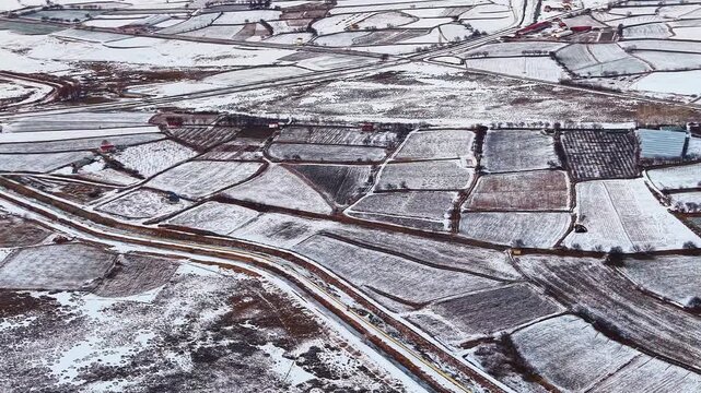 Drone aerial view of snow covered farmland stretching across a wide mountain valley. Patchwork fields, winding roads and winter terrain create a calm rural landscape beneath distant mountains.