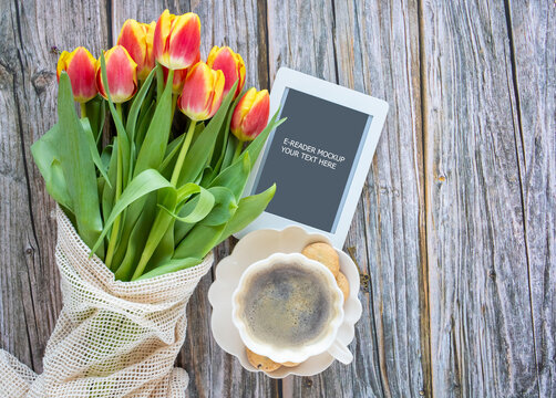 Cozy composition with cup of coffee, red-yellow tulips and e-reader with copy space on wooden background. Template for feminine blog social media.