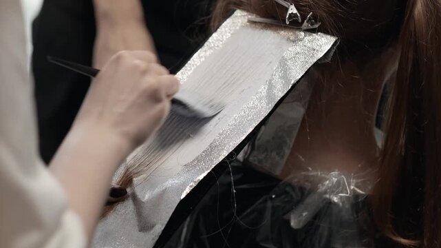 Professional hair colorist applying bleach to a section of hair on aluminum foil using a brush in a salon