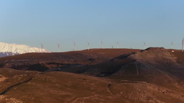 Aerial view of wind turbines standing tall against a backdrop of snow-capped mountains, contrasting with the brown and green tones of the landscape, Cocullo, Abruzzo, Italy.
