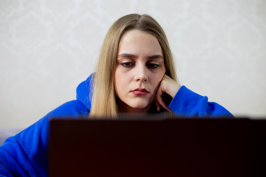Blonde girl forced to work remotely stares at laptop without interest. Unhappy employee stuck at home, no joy in online tasks. Sad remote worker concept