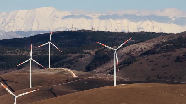 Aerial view of multiple wind turbines standing tall against a backdrop of snow-capped mountains and rolling hills, Cocullo, Abruzzo, Italy.