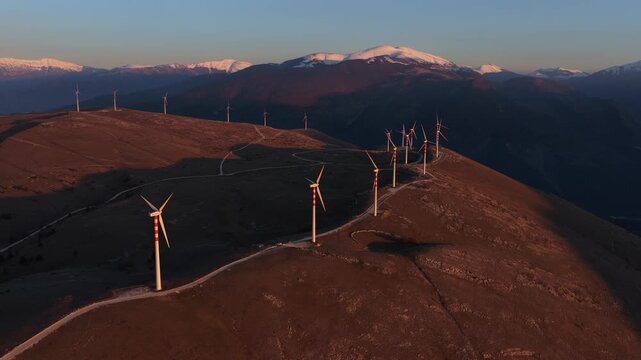 Aerial view of wind turbines standing tall on the mountain ridges with snow-capped mountains in the background, Cocullo, Abruzzo, Italy.