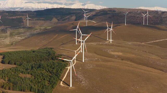 Aerial view of wind turbines standing tall on undulating hills, casting shadows on the ground, contrasted by a backdrop of snow-capped mountains, Cocullo, Abruzzo, Italy.