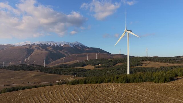 Aerial view of wind turbines contrasting against the snow-capped mountain and green trees, harnessing clean energy, Cocullo, Abruzzo, Italy.