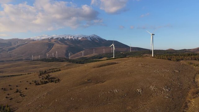 Aerial view of multiple wind turbines standing tall on brown grassy hills under a blue sky near snow capped mountains, Cocullo, Abruzzo, Italy.
