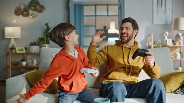 Father son gamers rejoicing win video game sitting home sofa hugging closeup. 