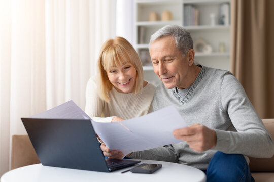 Senior couple reviewing documents together while sitting at table with laptop at home. Mature man and woman analyzing paperwork representing finance, retirement planning and household budgeting