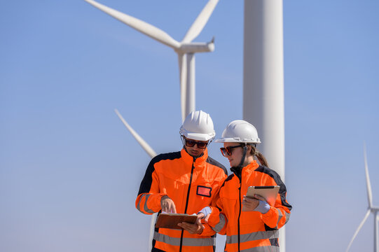Two wind energy engineers wearing safety helmets and high visibility jackets review technical data at wind farm for inspection and maintenance planning