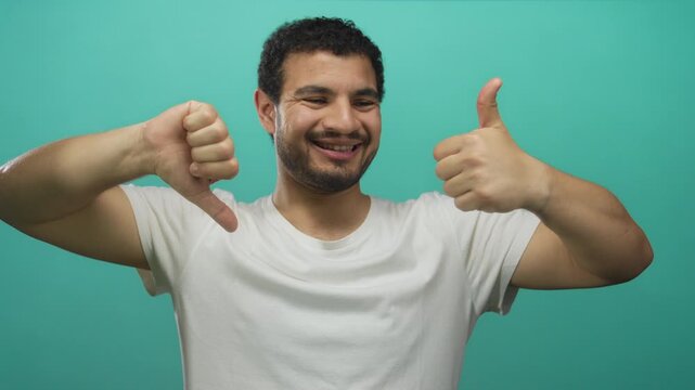 Man smiles broadly while showing thumb up on right hand and thumb down on left hand in aqua studio; indecision.