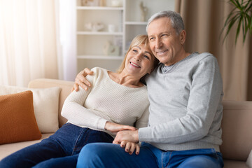 Obraz premium Senior couple sitting closely together on sofa at home looking away thoughtfully. Mature man and woman sharing quiet moment representing companionship, emotional support and retirement lifestyle