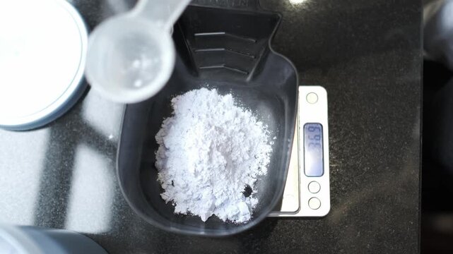 Hairstylist pouring white bleaching powder from a plastic scoop into a black mixing bowl on a digital scale. Top view