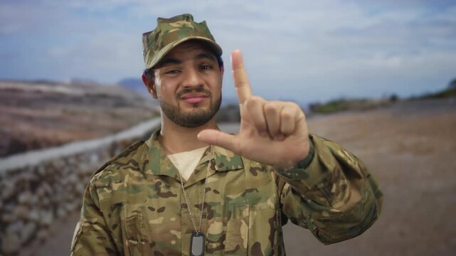 Man in camouflage uniform with military cap and dog tags points index finger at camera and holds l shaped hand gesture in street; pride duty.