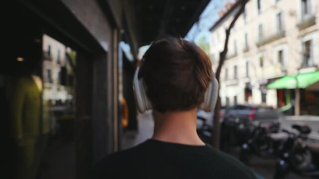 Rear view of a young man wearing modern white over-ear headphones, walking casually down a vibrant city street on a sunny day. He navigates past storefronts in Madrid, Spain