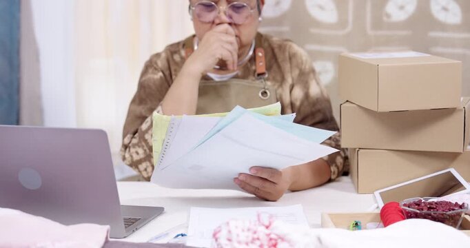 Senior artisan businesswoman thinks in slow motion, holds paperwork stack, checks laptop order status, reviews invoice totals, plans parcel shipping sequence inside home studio workspace. home studio