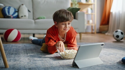 Smiling boy watching tablet cartoon eating popcorn lying apartment floor closeup © stockbusters