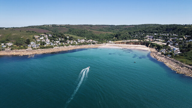 Aerial view of turquoise waters embrace the sandy beach nestled between rocky outcrops and verdant hills, a small boat leaving a white trail, Manche, Normandy, France.