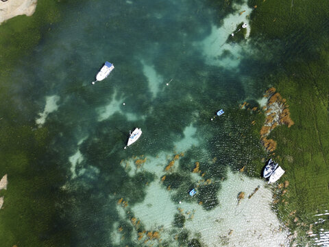 Aerial view of boats floating on shimmering turquoise waters, interspersed with patches of green seaweed, creating a mesmerizing mosaic of color and texture, Manche, Normandy, France.