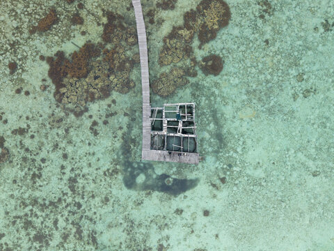 Aerial view of a weathered wooden pier extending into the turquoise sea near Cottage Legon Waru, contrasting with the submerged coral reefs, Karimunjawa, Central Java, Indonesia.