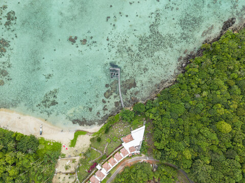 Aerial view of a vibrant coastline where emerald forests meet turquoise waters near a stilted structure, Cottage Legon Waru, Karimunjawa, Central Java, Indonesia.