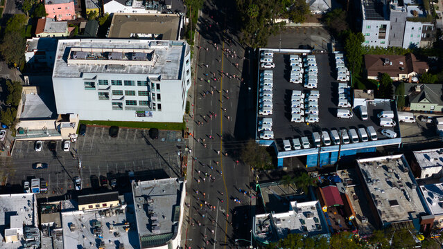 Aerial view of a vibrant parade flowing down a sun-drenched boulevard, framed by geometric buildings and rows of parked vehicles, West Hollywood, California, United States.