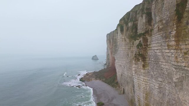 Aerial tracking pan of cliffs and sea stack along rocky coastline with waves lapping on pebble shore in misty morning