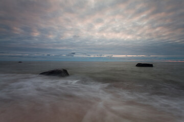 Fototapeta premium A tranquil Baltic Sea showing smooth, long-exposure waves flowing around large coastal rocks on a sandy beach beneath a pastel cloudy sunset, conveying calm, solitude and natural coastal beauty. 