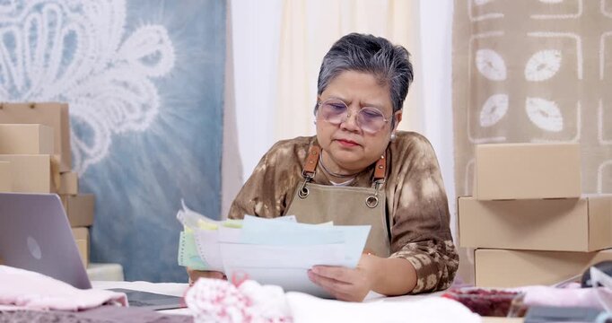 Senior asian entrepreneur speaks while reading paperwork, checks online order list on laptop, compares invoice with receipt, tracks parcel boxes, organizes shipping batch, studio dispatch continues.