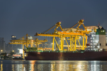 Container cargo ship at industrial port with cranes and city skyline at night.