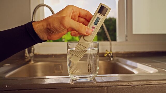 First Person View Of Hand Measuring Drinking Water With Digital Tester Showing Twenty Two Ppm In Glass On Kitchen Sink With Tropical Landscape Through Window. Clean Drinking Water Quality