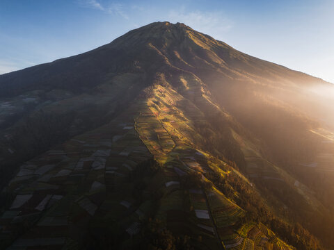 Aerial view of a towering mountain peak pierced by the sun's golden rays, casting long shadows across the cultivated terraces below, Nepal van Java, Jawa Tengah, Indonesia.