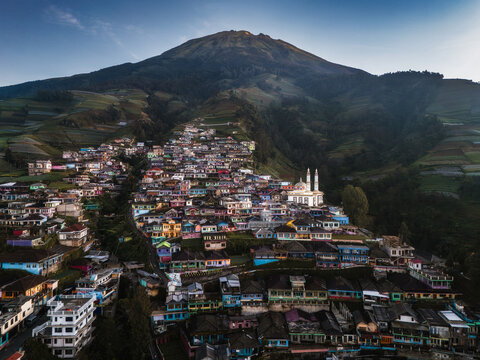 Aerial view of colorful houses cascading down the hillside towards a bright white mosque, with a majestic mountain looming in the background, Nepal van Java, Jawa Tengah, Indonesia.