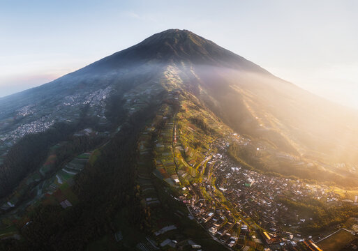 Aerial view of a majestic mountain peak piercing through the clouds, casting long shadows over the terraced fields and clustered houses below, Nepal van Java, Jawa Tengah, Indonesia.
