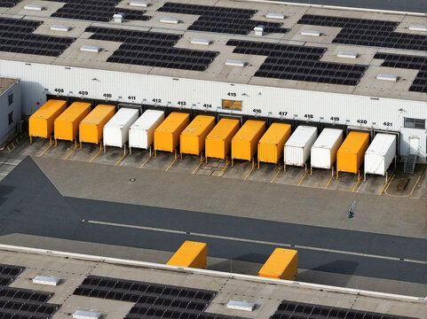 Aerial view of rows of white and yellow trucks backed up against the loading docks of a warehouse with solar panels on its roof, Unspecified City, Unspecified Country.