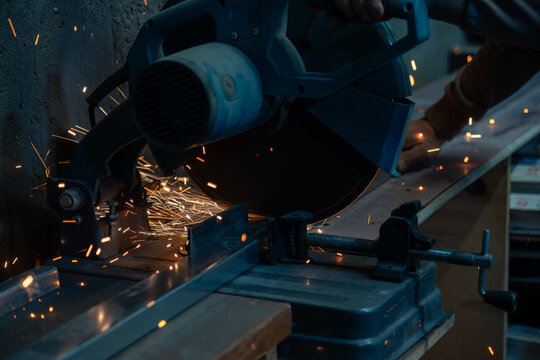 Worker cutting metal profile with electric miter saw, flying sparks in industrial metal fabrication workshop.