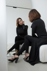 Obraz premium Stylish young woman sitting on a white ottoman, admiring her look in the mirror. High-fashion minimalist aesthetic featuring a sleek black outfit and trendy eyewear in a bright studio.