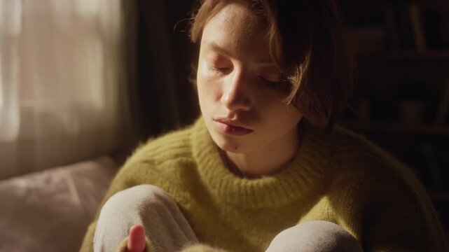 Tilt up shot of melancholic young adult woman dressed in long sleeved sweater sitting on bed in sunlight fidgeting with ring while thinking anxiously in bedroom