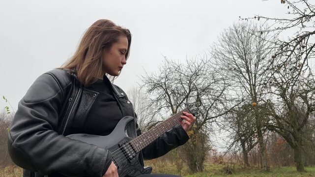 young female rock musician in a leather jacket plays an electric guitar outdoors