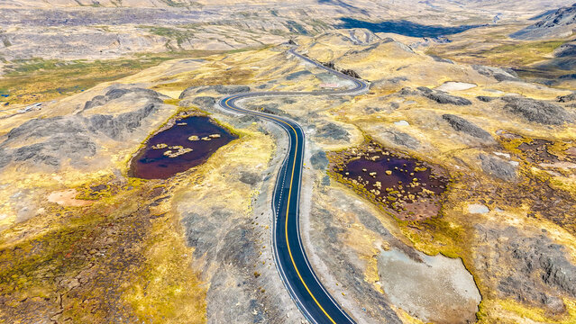 Aerial view of a winding road cutting through the rugged landscape, punctuated by dark pools reflecting the sky's hue, Pajchanta, Qosqo, Peru.
