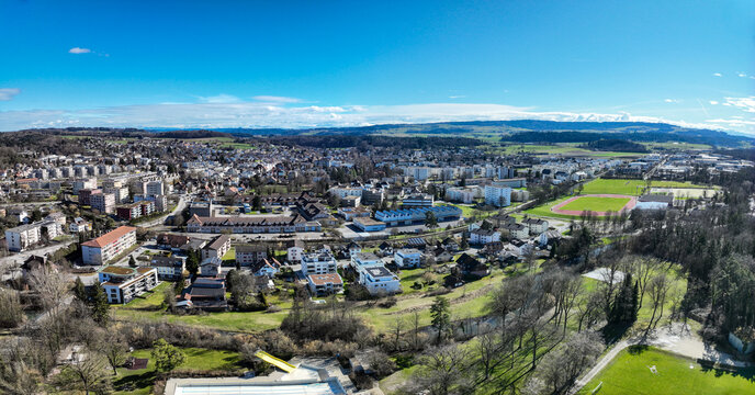 Aerial view of a vibrant townscape with residential buildings, interspersed with green spaces, under a clear blue sky, Lyss, Canton of Bern, Switzerland.