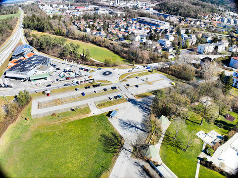 Aerial view of a roundabout and parking area with cars nestled between lush green fields and a cityscape under a bright sky, Lyss, Canton of Bern, Switzerland.